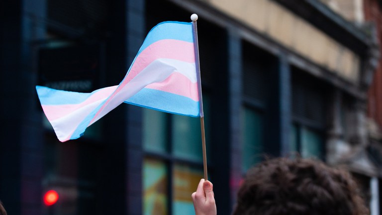 Uma mão segura uma bandeira trans. A bandeira tem cinco faixas de cores, na seguinte ordem: azul, rosa, branco, rosa e azul