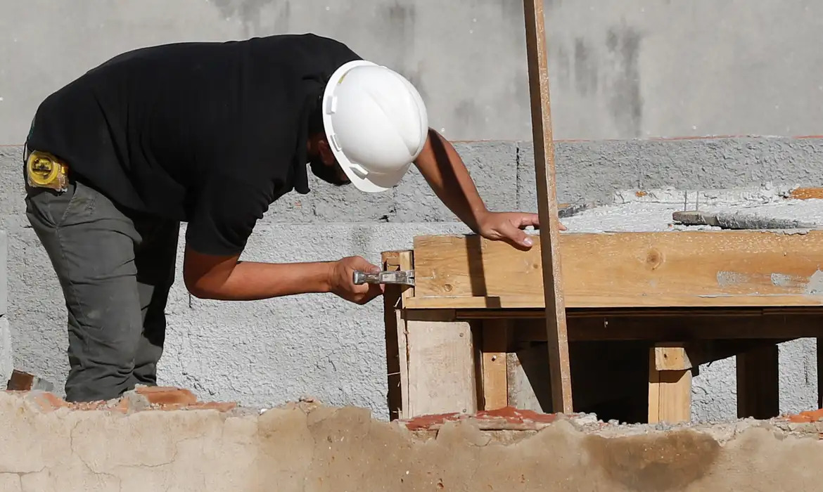 Um trabalhador da construção civil martela uma estrutura de madeira dentro do que parece ser um prédio em construção