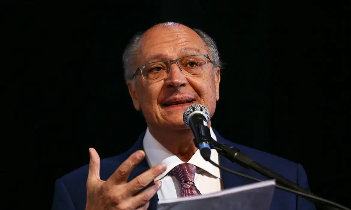 Geraldo Alckmin é um homem idoso, com topo da cabeça calva e cabelos brancos nas laterais. Veste um par de óculos e uma roupa social, e parece discursar diante de um microfone