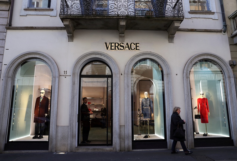 Pessoa caminha em frente a uma loja com vitrines para a rua. É possível ler no letreiro o nome da grife "Versace"