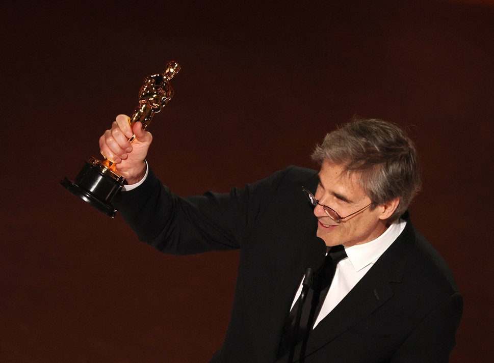 Walter Salles fala diante do microfone no teatro da cerimônica do Oscar enquanto exibe o troféu com a mão erguida acima da cabeça