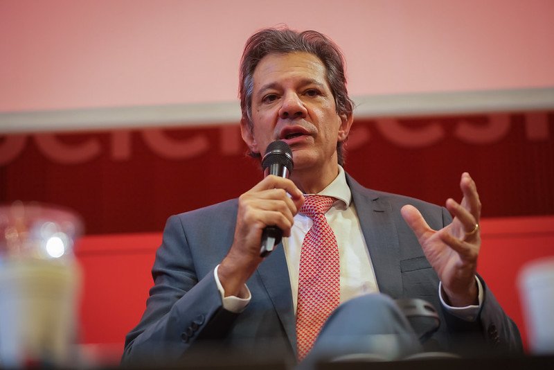 Fotografia do ministro da Fazenda, Fernando Haddad. Ele segura um microfone próximo da boca com uma mão e gesticula com a outra
