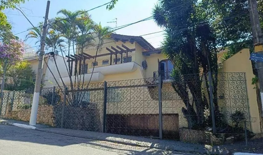 Fotografia da casa com maior lance inicial no leilão. É uma casa com vários andares, amarela, com cerca de ferro vazado e árvores no quintal