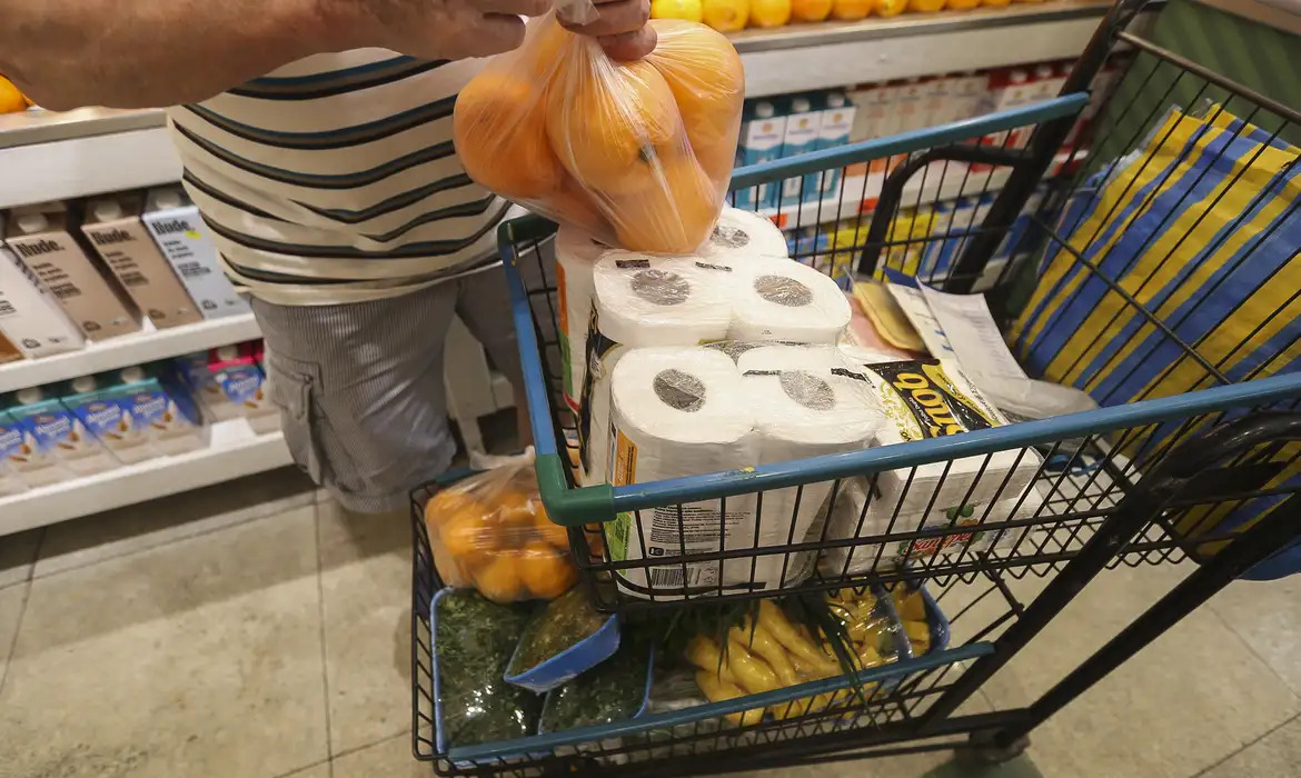 Em um carrinho de supermercado, estão alimentos que compõe a cesta básica, além de frutas e itens de higiene como papel higiênico