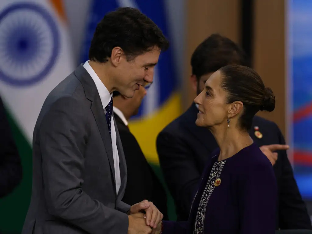 Justin Trudeau e Claudia Sheinbaum, presidentes do Canadá e do México respectivamente, falam um de frente para o outro enquanto apertam as mãos. Os dois sorriem