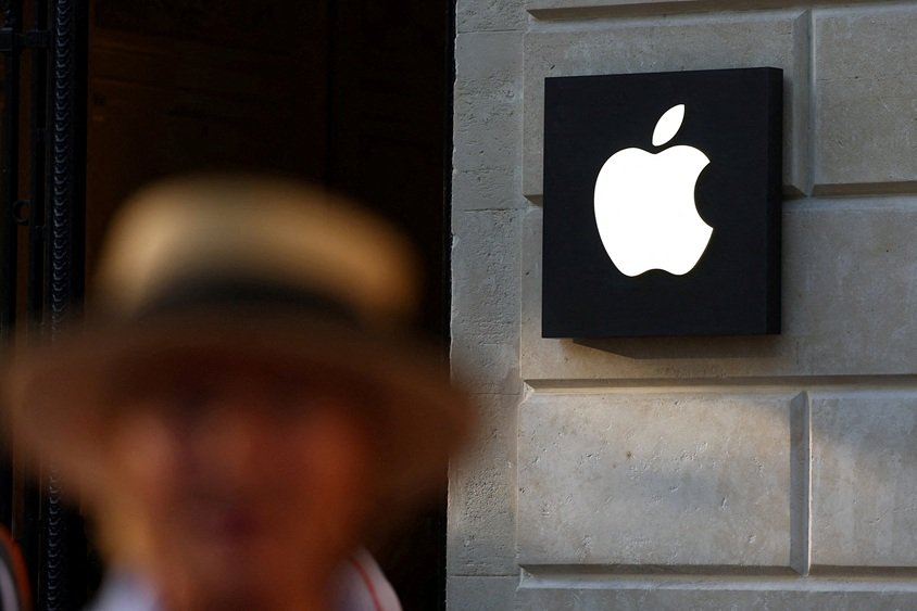 Na fotografia, um homem aparece desfocado em primeiro plano e, ao fundo, vemos o logo da Apple