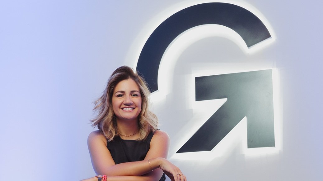 Mariana Dias é uma mulher loira e jovem, que aparece sorrindo em frente a uma parede com o logo da Gupy