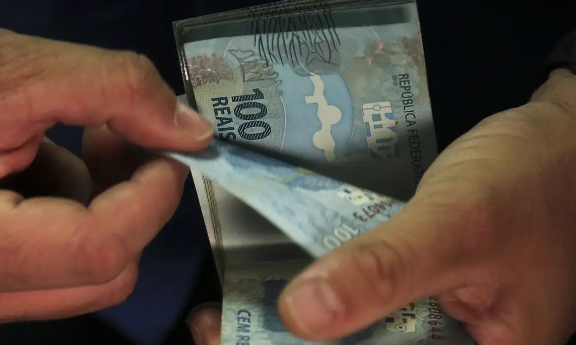 Fotografia de duas mãos a contar notas de 100 para ilustrar matérias sobre investimentos no PIB