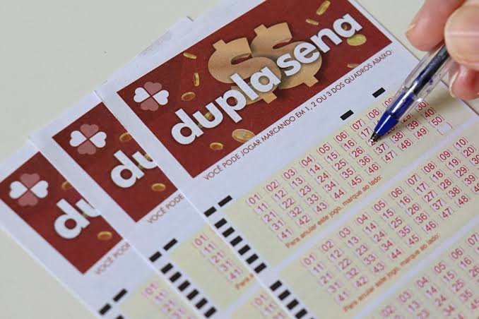 Mão de pessoa segurando um bilhete de loteria Dupla Sena, com as dezenas e o logo da Caixa visíveis, simbolizando o sorteio.