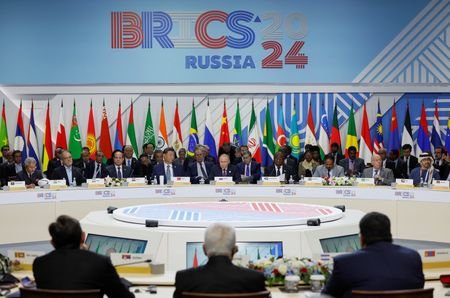 Em torno de uma mesa redonda, estão reunidos diversos chefes de estado. Ao fundo, vê-se várias bandeiras, uma ao lado da outra, e o logo do Brics.